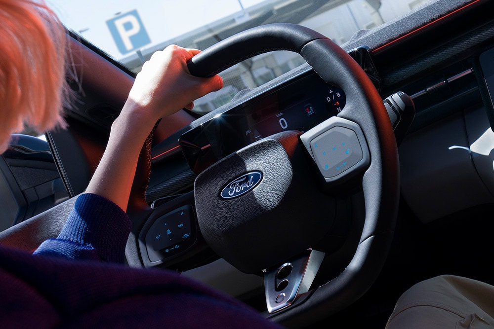 Nahaufnahme einer Frau, die das Lenkrad in einem geparkten Ford Capri hält, wobei auch das digitale Kombiinstrument zu sehen ist.