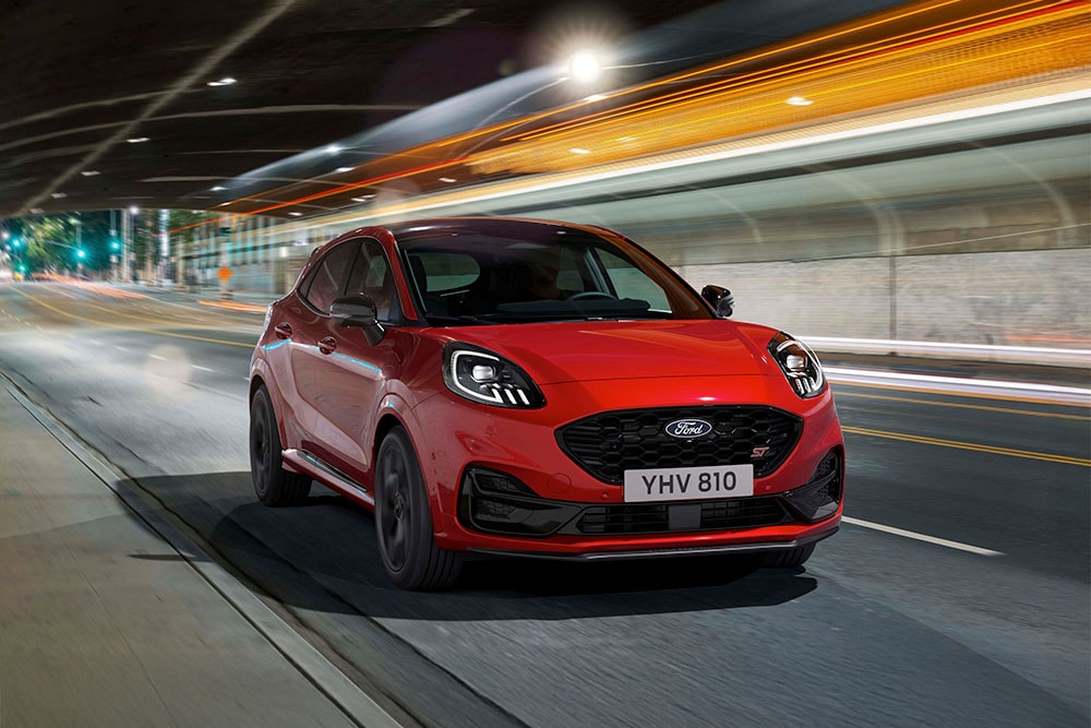 Ein sportlicher SUV des Modells Ford Puma ST fährt nachts auf einer hell erleuchteten Stadtstrasse.