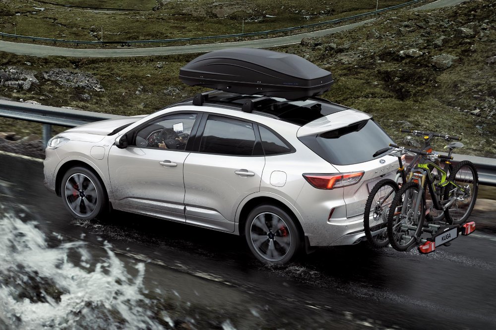 Ford Kuga in silber fahrend auf Strasse in den Bergen mit Heckträger und Fahrrädern. Dreiviertelansicht hinten.