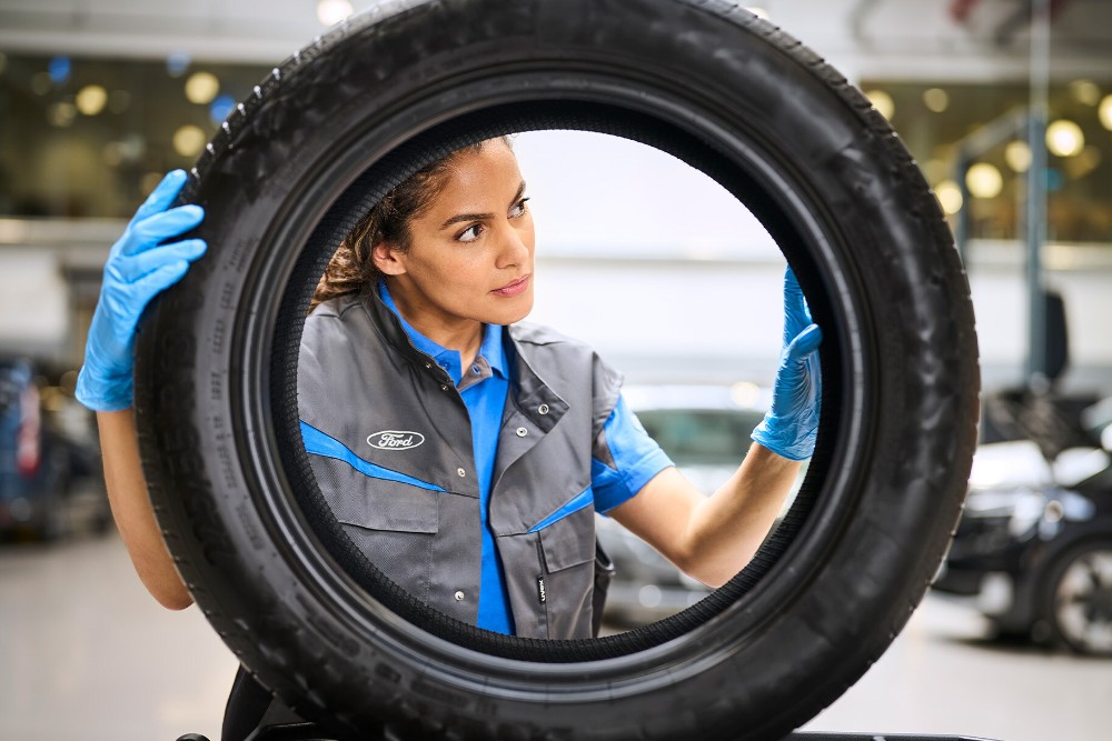 Eine Ford Fachkraft mit blauen Handschuhen prüft einen hochgehaltenen Reifen in der Werkstatt. 