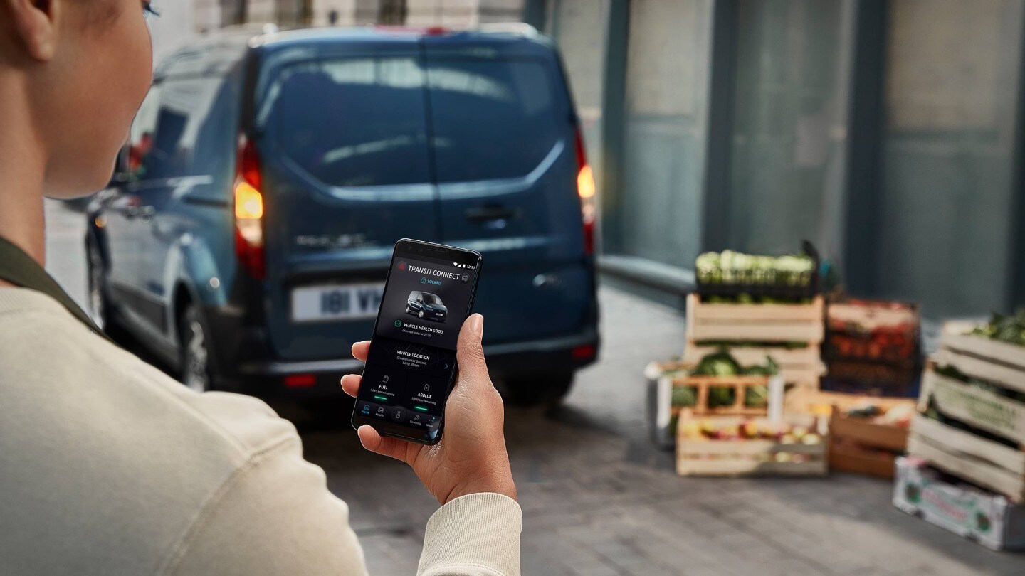 FordPass App – Frau hält Mobiltelefon und Ford Transit Connect in Blau Heckansicht parkt in Lagerhalle