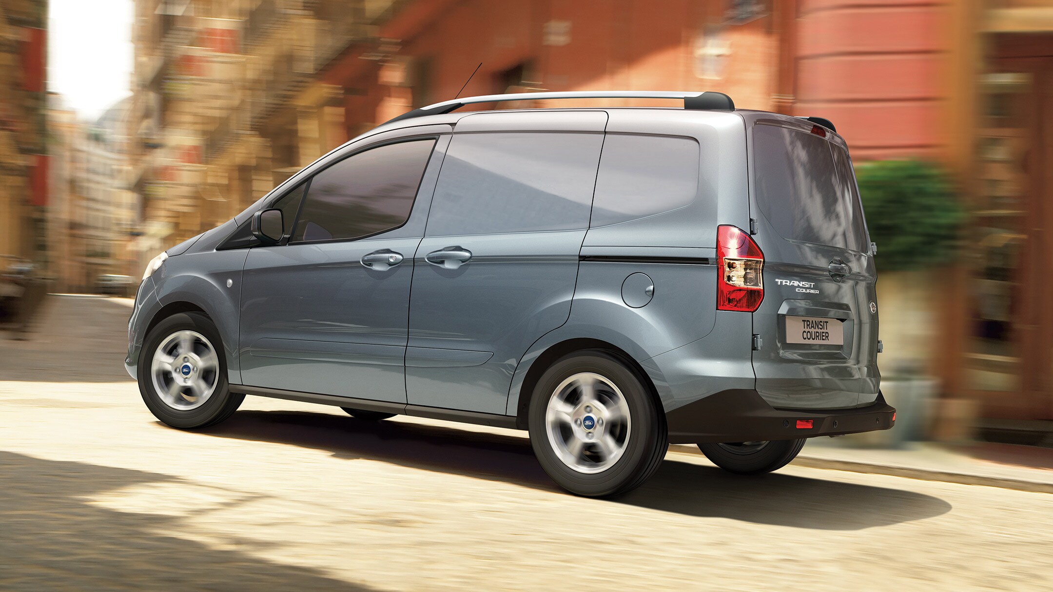 Ford Transit Courier in Grau Seitenansicht fährt durch enge Strasse in der Innenstadt