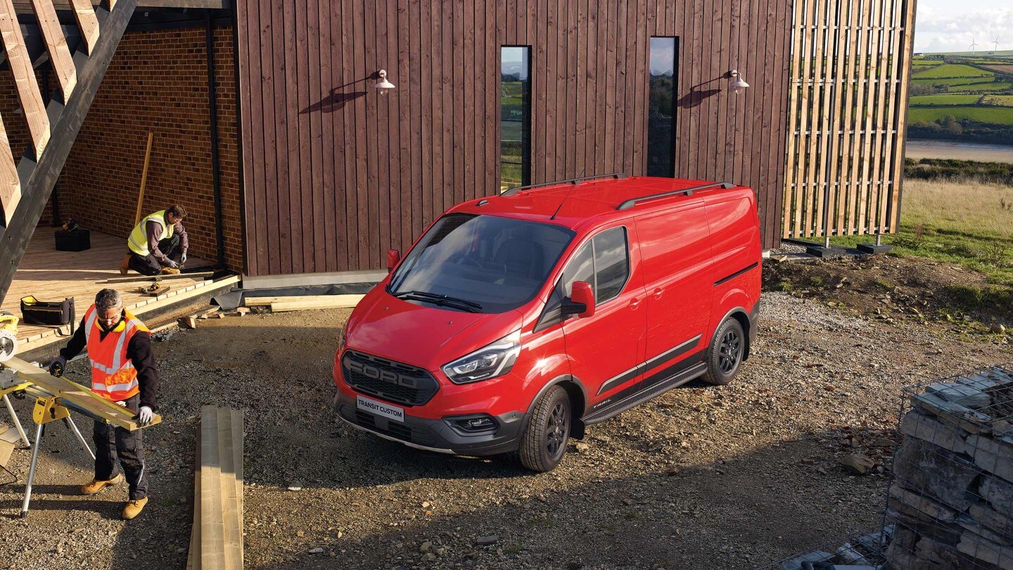 Ford Transit Custom Trail Rot ¾-Seitenansicht parkt auf Baustelle vor Holzhaus