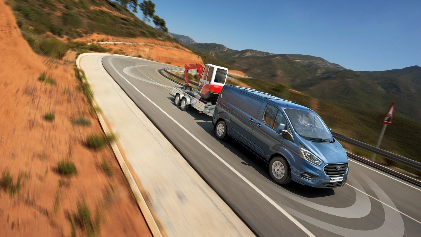 Ford Transit Custom in Blau ¾-Frontansicht mit Anhänger fährt bergab auf Landstrasse