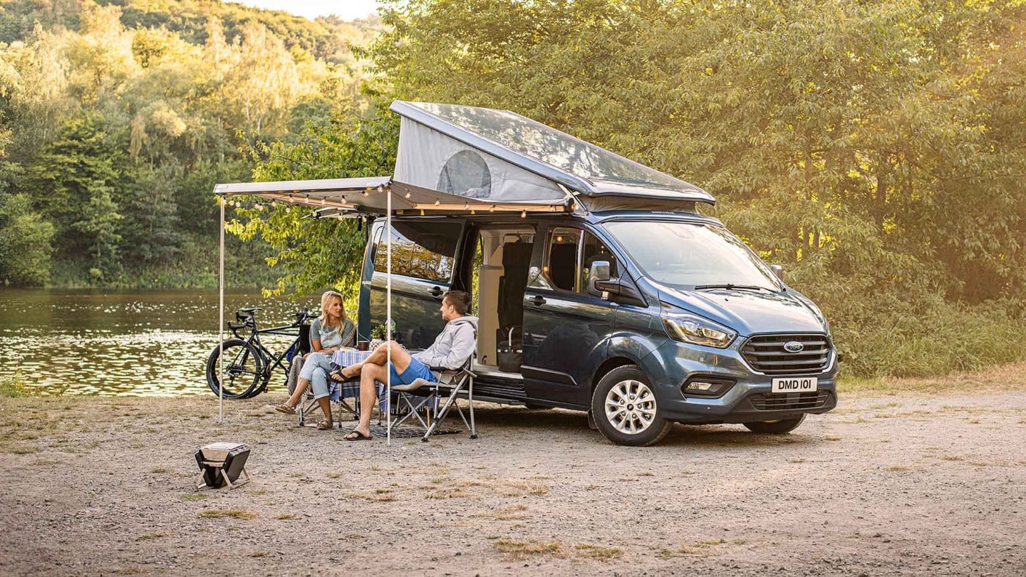 Ford Transit Custom Nugget in Blau mit Aufstelldach in der Seitenansicht mit unter Vordach sitzenden Personen parkt an Seeufer