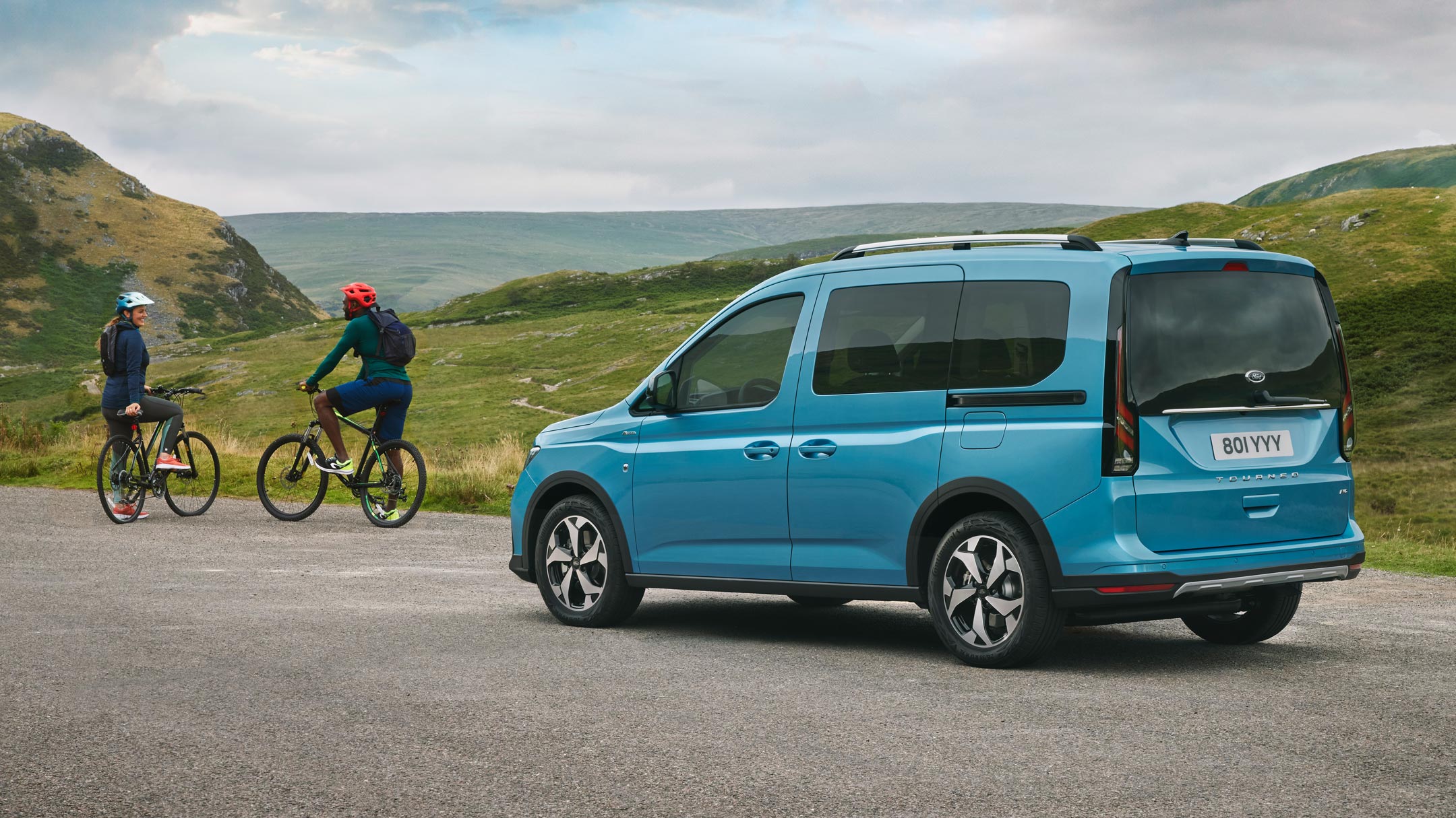 Ford Tourneo Connect in Hellblau ¾-Heckansicht steht auf Landstrasse, davor stehen zwei Fahrradfahrer