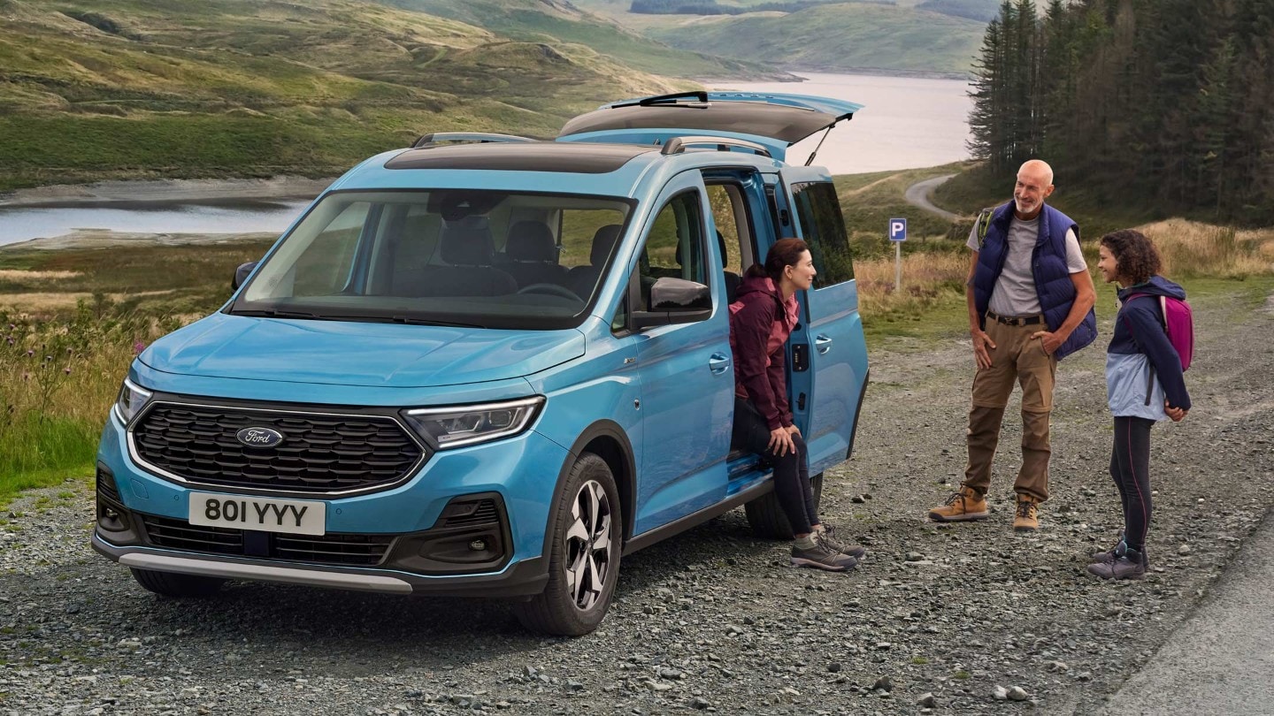 Ford Tourneo Connect Connect in Hellblau ¾-Frontansicht mit geöffneter Schiebetür parkt auf Schotterweg. Frau sitzt im Fahrzeug, Mann und Mädchen stehen daneben