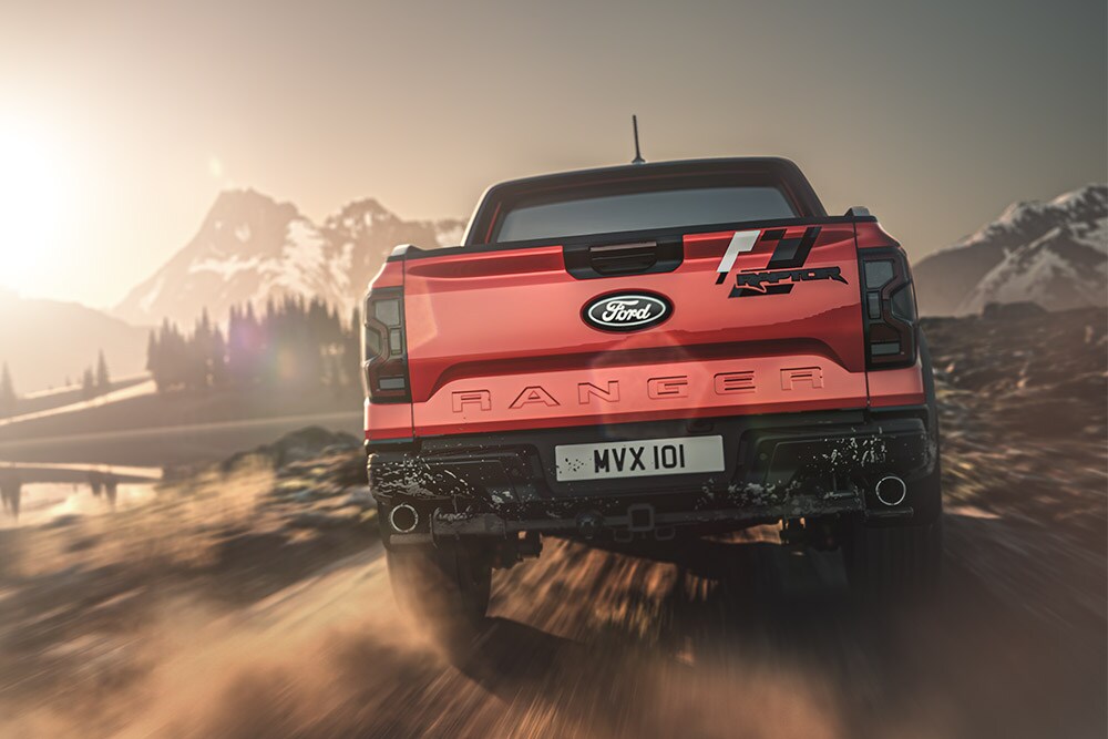 Heckansicht eines roten Ford Ranger Raptors, der auf einem Feldweg fährt, die Berge im Hintergrund.
