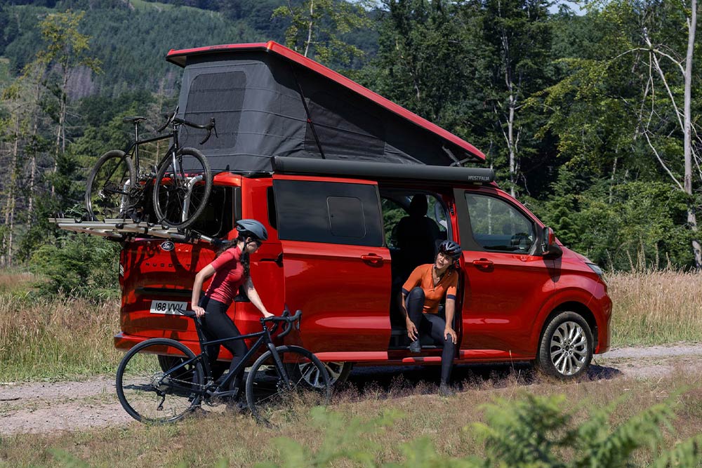 Personen laden Velos vom Fahrradträger eines roten Ford Nugget ab.
