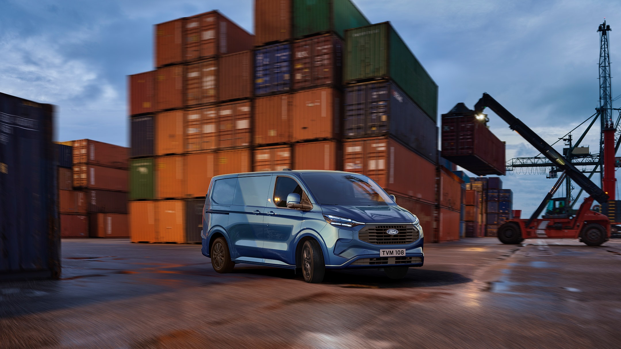Blauer Ford Transit Custom Plug-In Hybrid in einem Containerhafen vor gestapelten Überseecontainern.