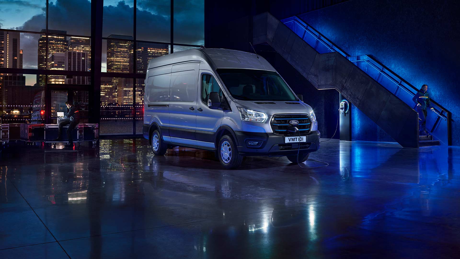 Silberner Ford E-Transit Elektro-Van , steht in einer Halle, durch die grossen Fenster ist die Skyline der Stadt zu sehen. 