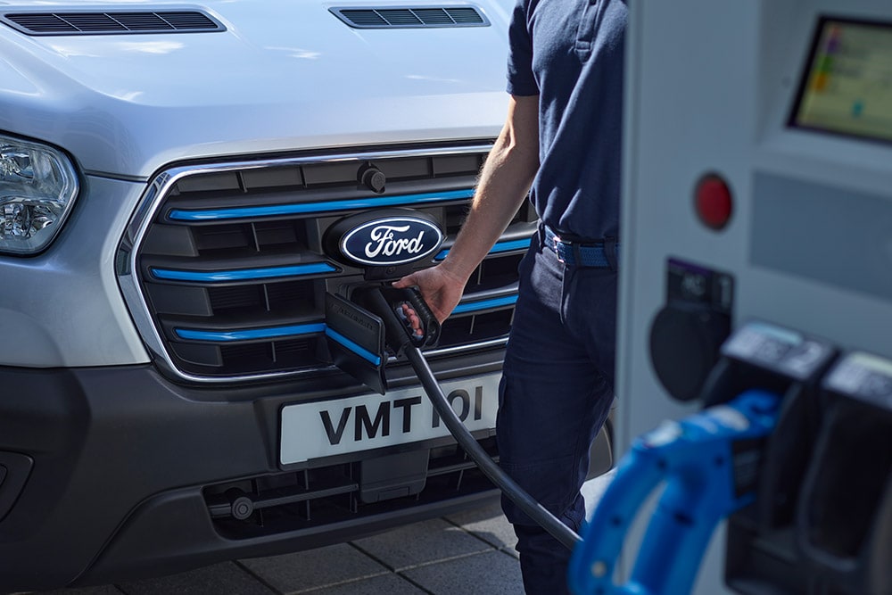 Nahaufnahme einer Person, die ein Ladekabel an einen silbernen Ford  E-Transit Elektro-Van anschliesst.