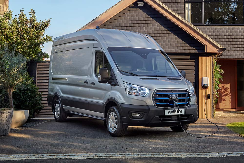 Silberner Ford E-Transit Van, geparkt in einer Einfahrt neben einem Haus, angeschlossen an eine Ladestation.