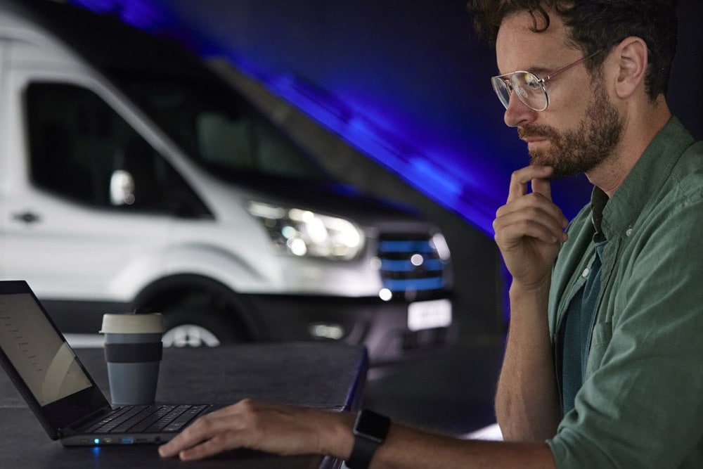 Ein Mann, der an einem Laptop arbeitet, im Hintergrund ein Ford E-Transit-Transporter.