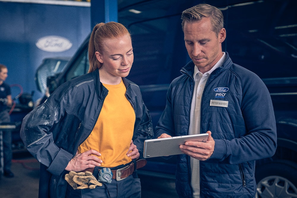 Zwei Mechaniker in einer Werkstatt, die vor einem Ford E-Transit-Transporter auf ein Tablet blicken.