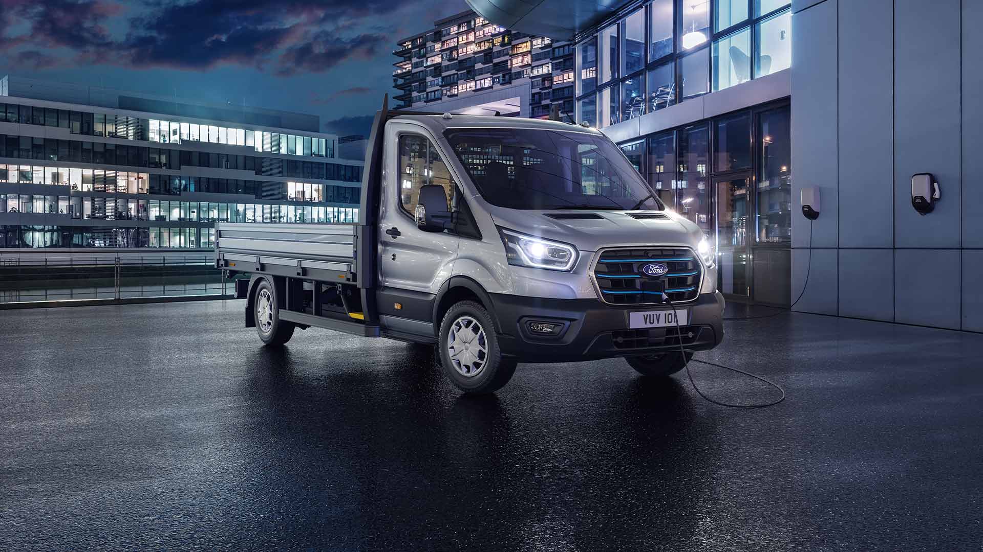 Silberner Ford Transit Fahrgestell mit Fahrerhaus, der nachts vor einem modernen Gebäude geparkt ist.