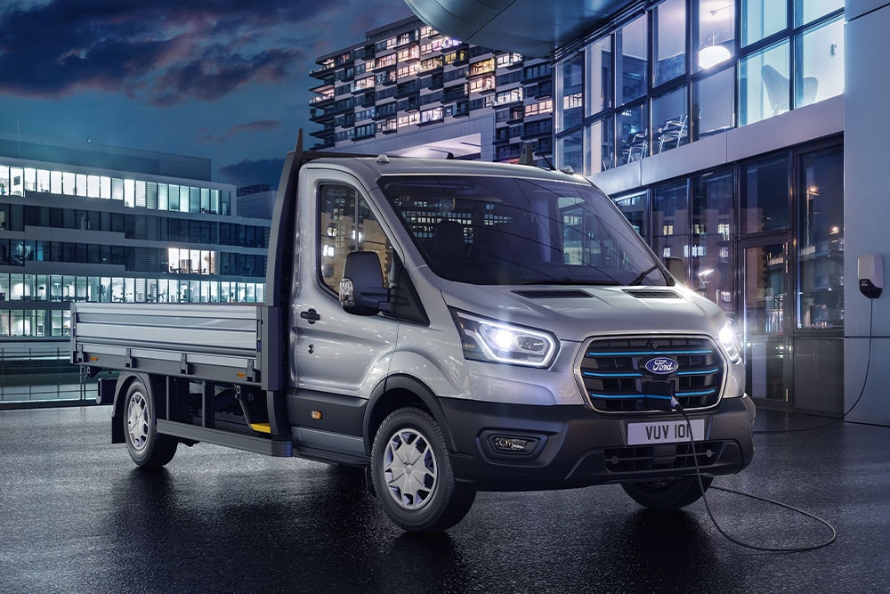 Silberner Ford Transit Fahrgestell mit Fahrerhaus, der nachts vor einem modernen Gebäude geparkt ist.
