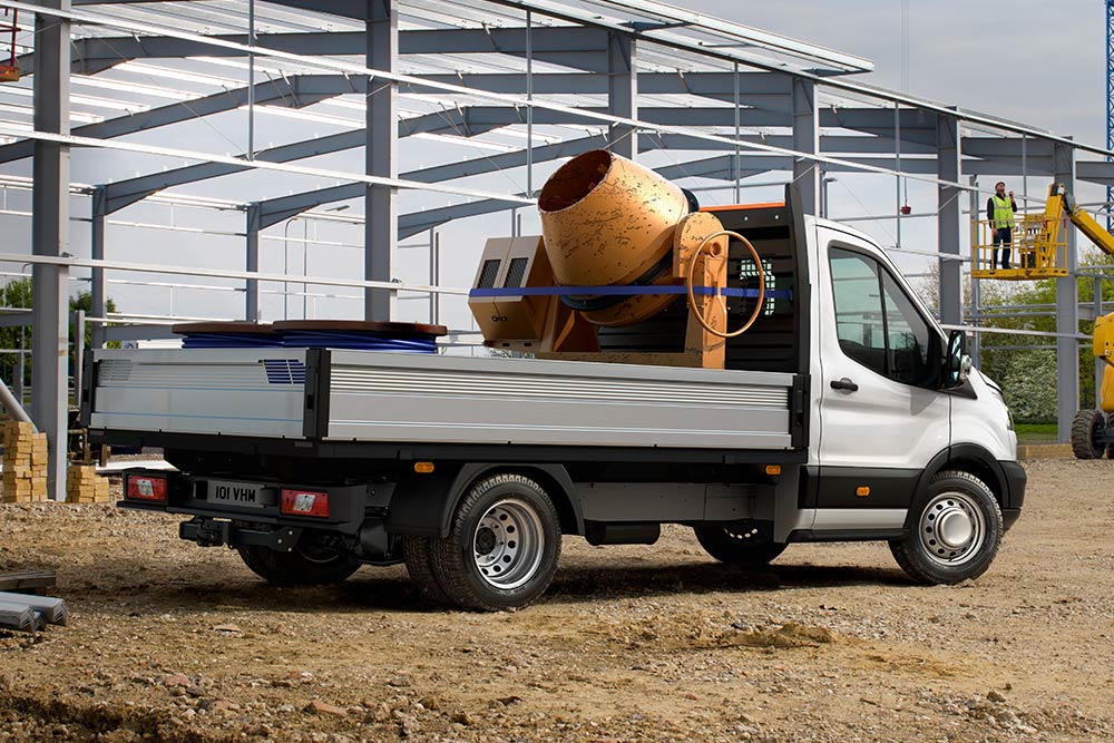 Ford Transit mit Fahrgestell und Fahrerhaus, der auf der Ladefläche einen Betonmischer transportiert und auf einer Baustelle geparkt ist. 