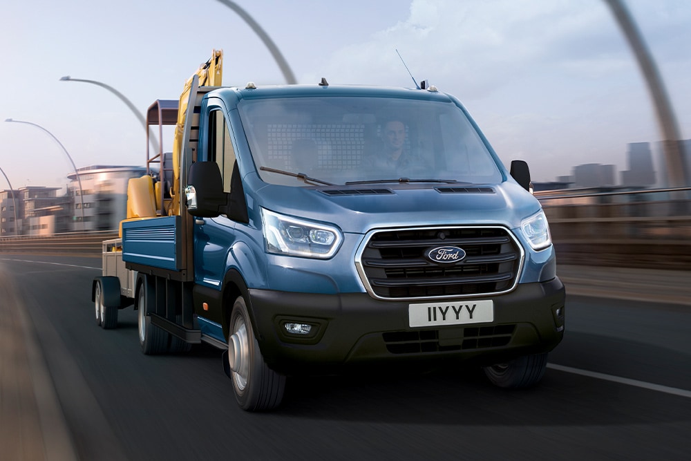 Frontansicht des Kühlergrills eines Ford Transit Fahrgestells mit Fahrerhaus mit Bagger während der Fahrt entlang einer Stadtstrasse. 