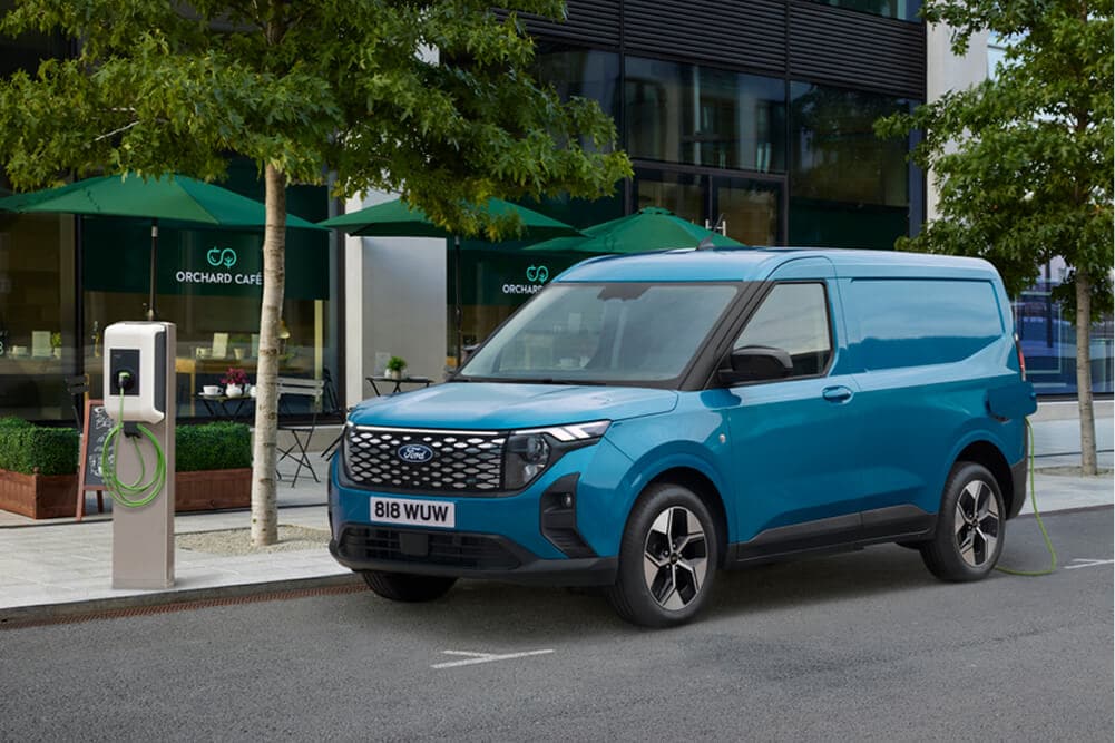 Blauer Ford E-Transit Courier im ¾-Frontansicht, parkiert neben einer Ladestation