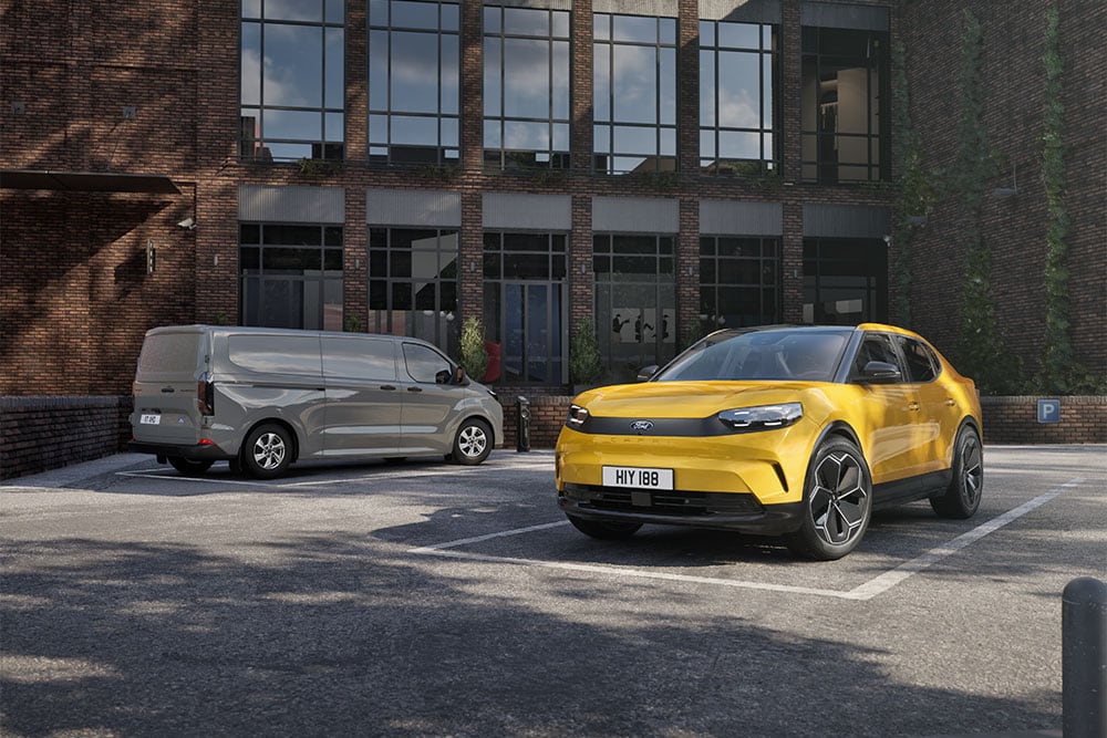  Ein grauer Ford-Transporter und ein gelber Capri, die vor einem modernen Backsteingebäude geparkt sind.