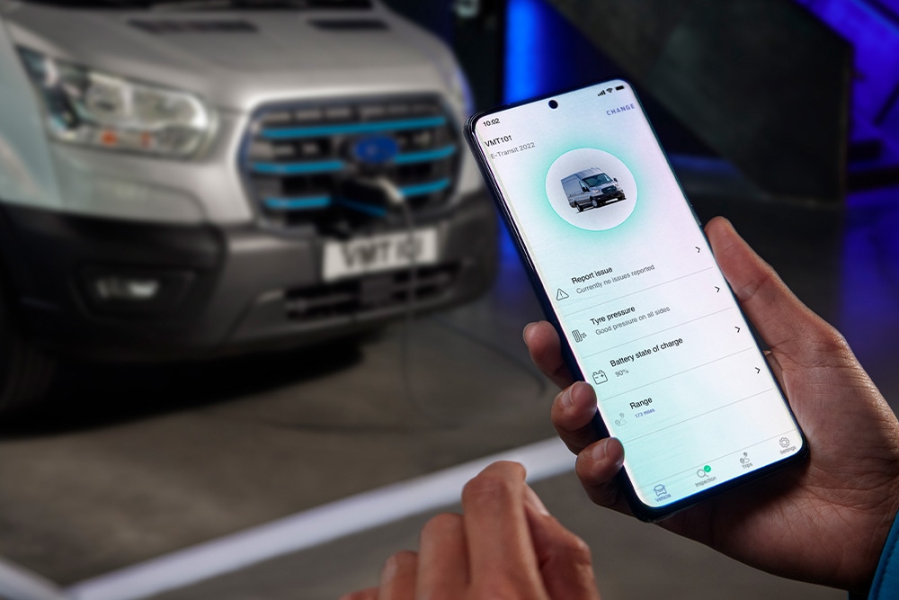 Eine Person hält ein Smartphone mit einer Ford App, während ein Ford E-Transit Custom im Hintergrund lädt.
