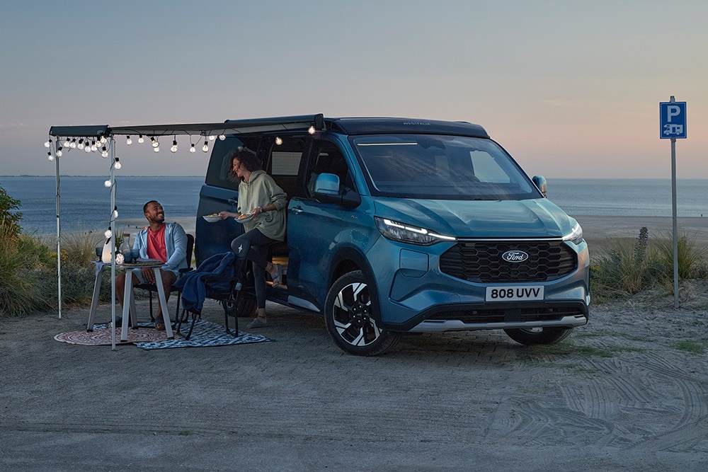 Zwei Camper entspannen sich neben einem blauen Ford Pro Transit Custom Nugget, ein Parkplatz am Strand im Hintergrund.