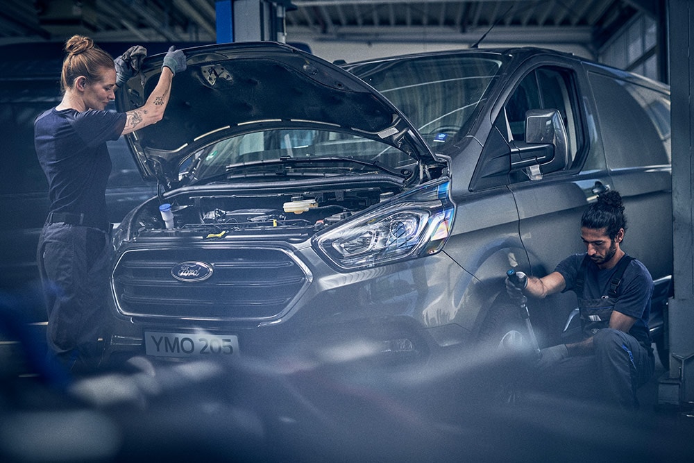 Zwei Ford Techniker reparieren einen Transit Custom in der Werkstatt. Einer am offenen Motor, der andere am Rad.