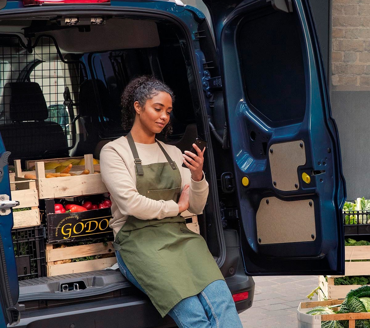 Händlerin schaut auf ihr Handy und lehnt an der Ladefläche eines Ford Transit Couriers