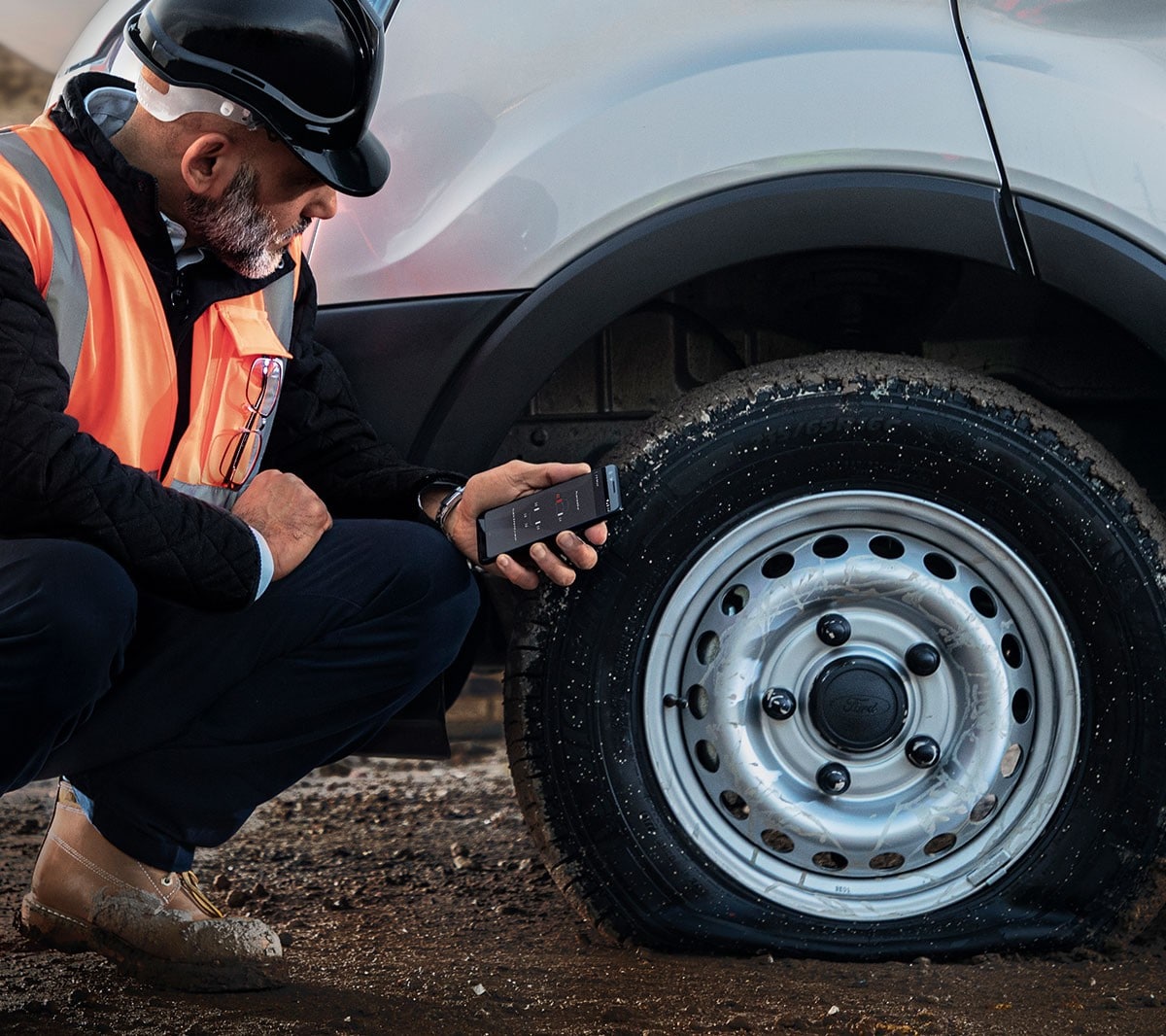 Arbeiter mit Bauhelm und Warnweste kniet neben einem Fahrzeug mit plattem Reifen und hält ein mobiles Telefon in der Hand