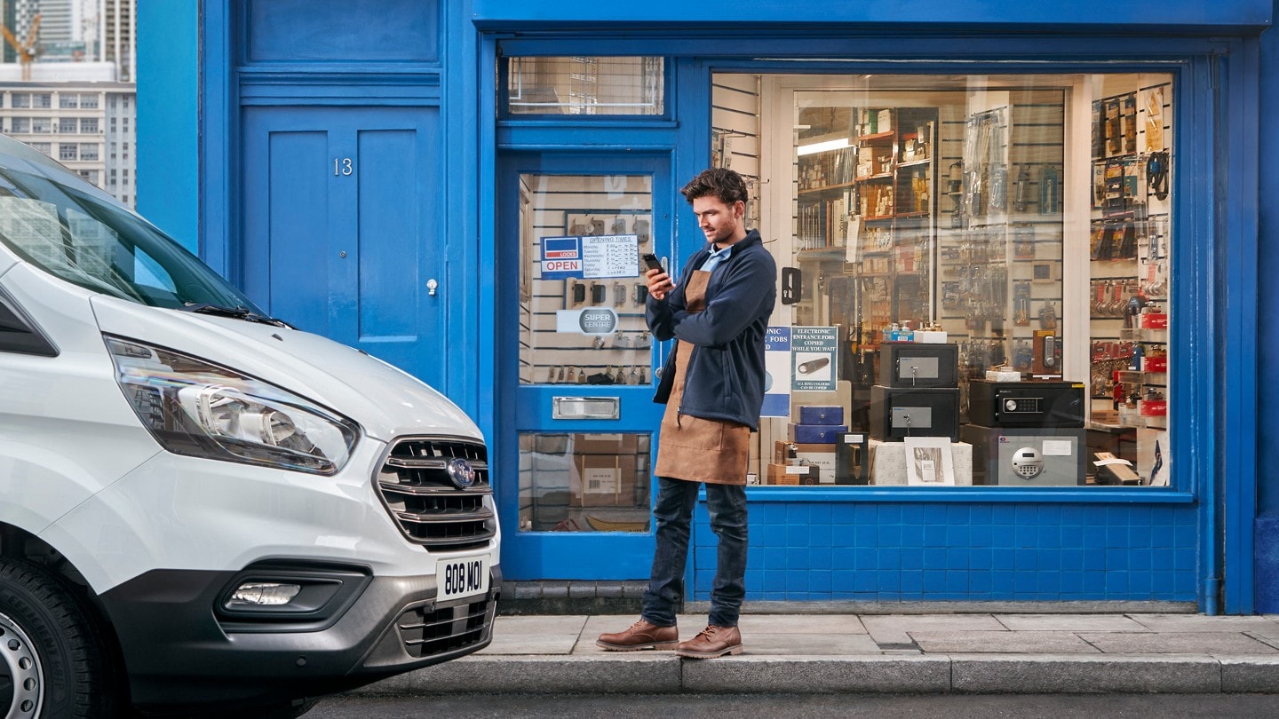 Mann steht vor einem Geschäft neben einem Firmenfahrzeug mit mobilem Telefon in der Hand 