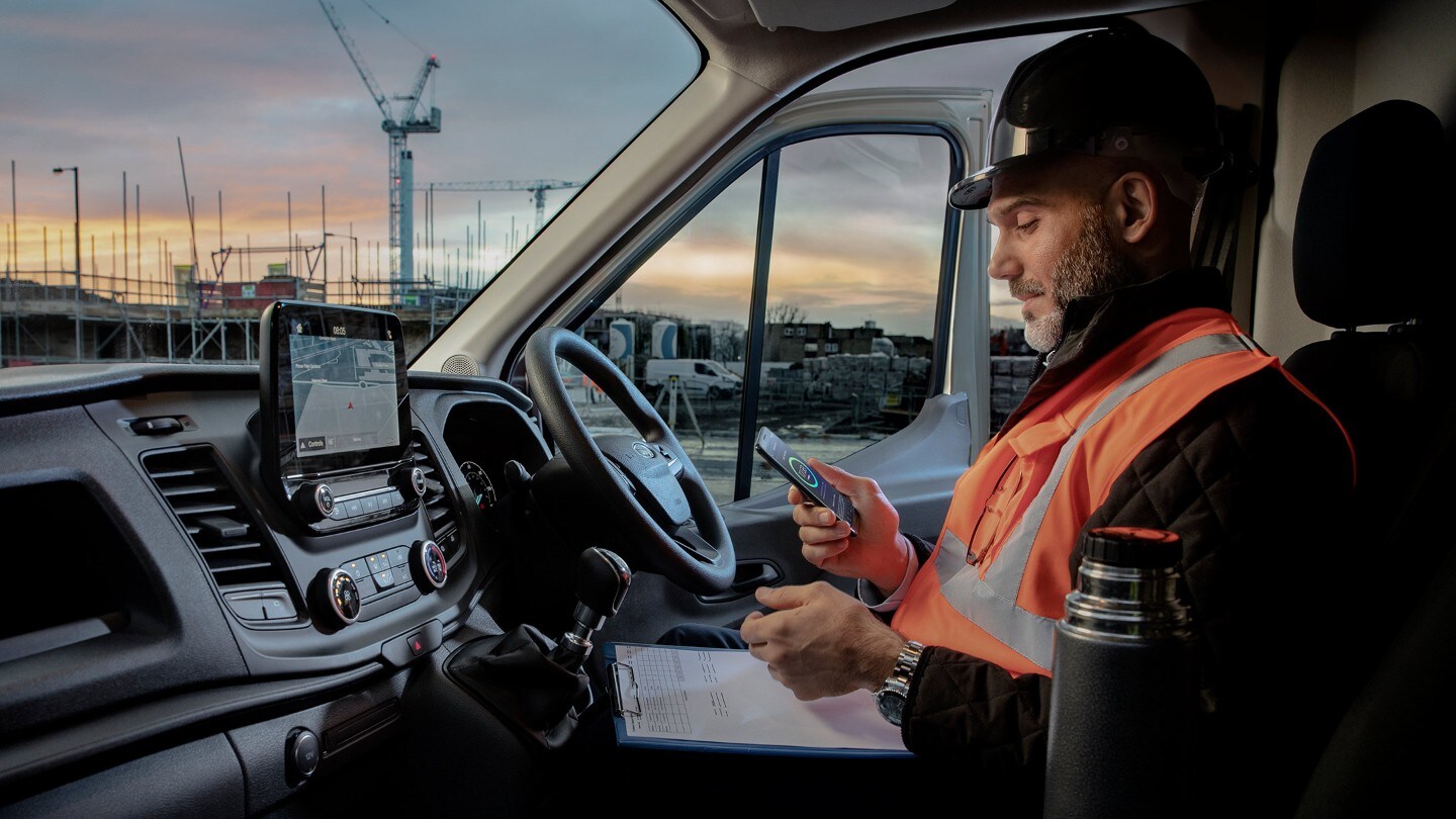 Arbeiter mit Bauhelm sitzt hinter dem Steuer eines Fahrzeugs und schaut auf ein mobiles Telefon