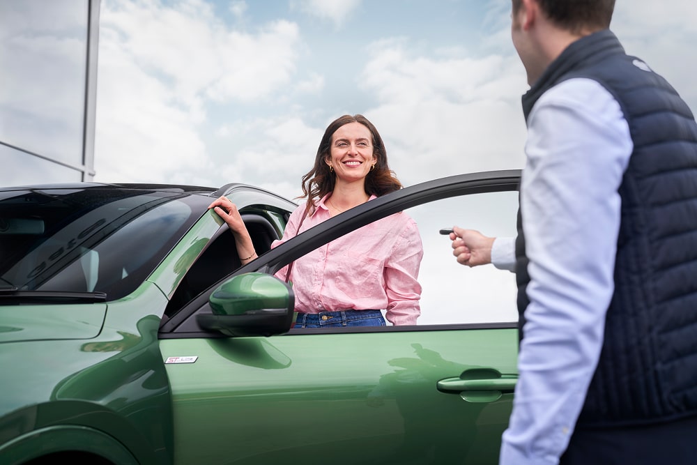 Eine Frau in einem rosa Hemd lächelt neben einem grünen Auto, während ein Ford-Händler ihre Autoschlüssel überreicht.
