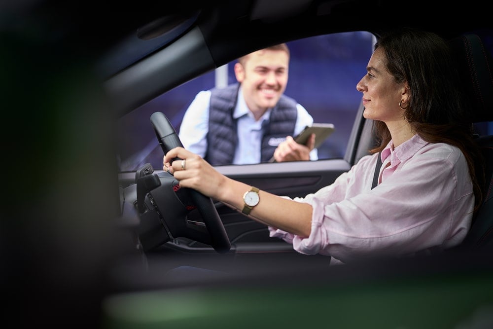 Eine Frau in einem geparkten grünen Auto lächelt einen Ford-Händler vor dem Fenster an.