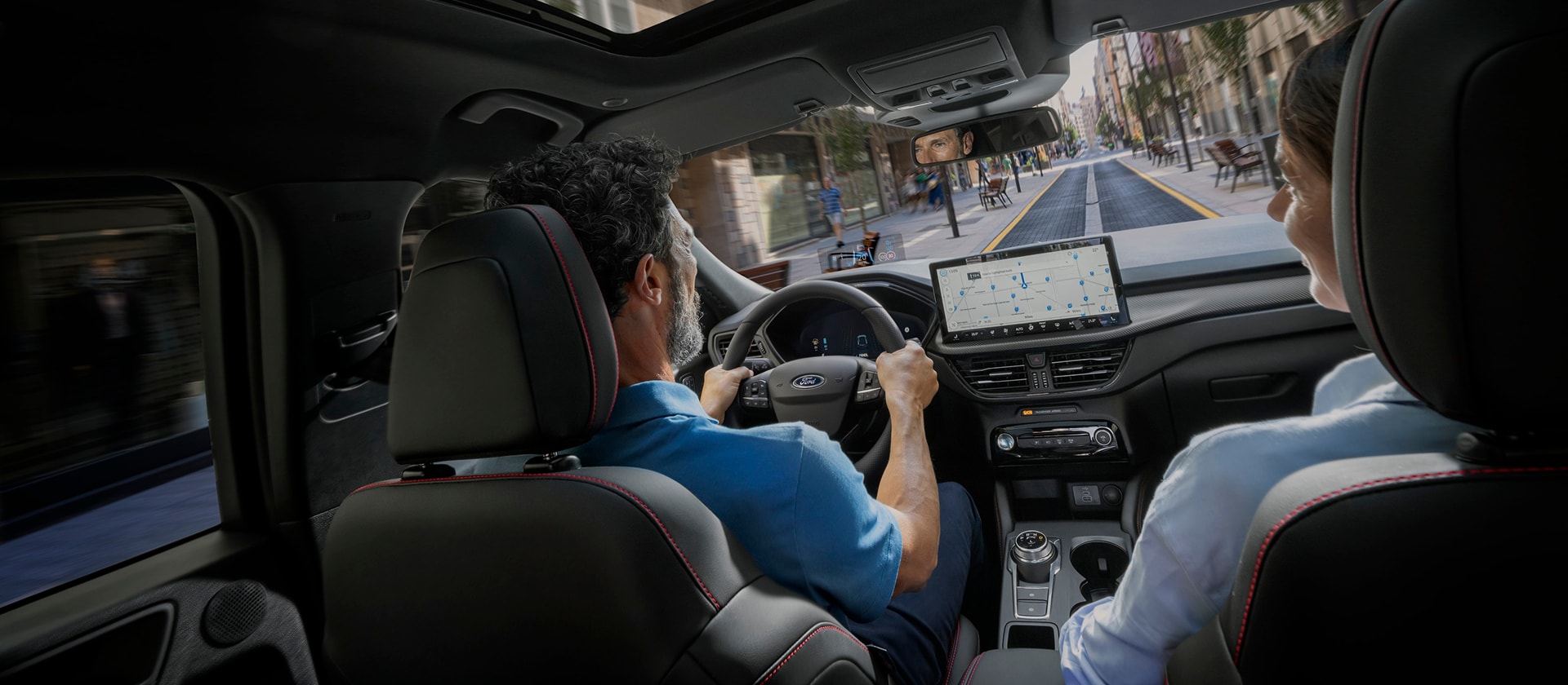 Conducente e passeggera anteriore di una Ford usano il SYNC sul display digitale per la navigazione urbana. 